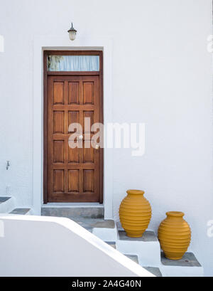 a wooden panelled door with two mustard coloured colored pots on steps against white wash concrete wall in Greece Stock Photo