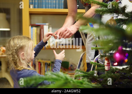Adorable little girl and her grandma decorating the Christmas tree with colorful glass baubles. Trimming the Christmas tree. Celebrating Xmas at home. Stock Photo