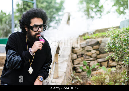 Vape man. Outdoor portrait of a young brutal white guy with large beard and in sunglasses letting puffs out of steam from an electronic cigarette opposite the old wooden fence in the village. Vaping. Stock Photo