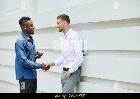 Cheerful male business colleagues shaking hands and greeting each other outside the office building Stock Photo