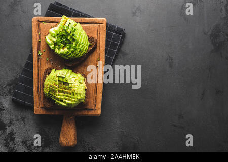 Two avocado sandwiches with rye bread on wooden cutting board, black concrete background. Top view with copy space. Clean eating, healthy vegan vegeta Stock Photo