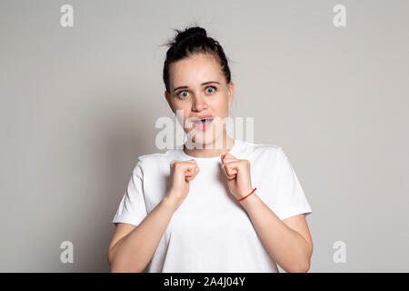 A pretty young woman in anticipation of a surprise emotionally and excitedly clenched her hands into fists, and looks into the camera with wide open e Stock Photo