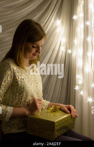 Portrait of a beautiful young white woman holding a christmas present in her hands in front of an illuminated star shaped christmas chain of lights Stock Photo