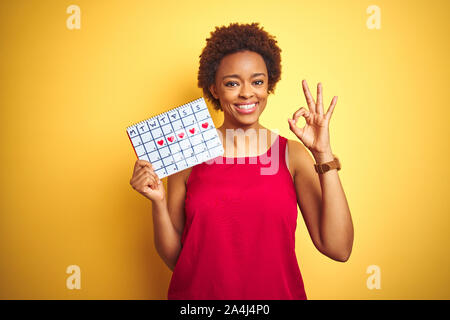 Young african american woman holding menstruation calendar over isolated yellow background doing ok sign with fingers, excellent symbol Stock Photo