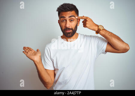 Young indian man wearing t-shirt standing over isolated white background confused and annoyed with open palm showing copy space and pointing finger to Stock Photo