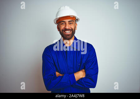 Handsome indian worker man wearing uniform and helmet over isolated white background happy face smiling with crossed arms looking at the camera. Posit Stock Photo