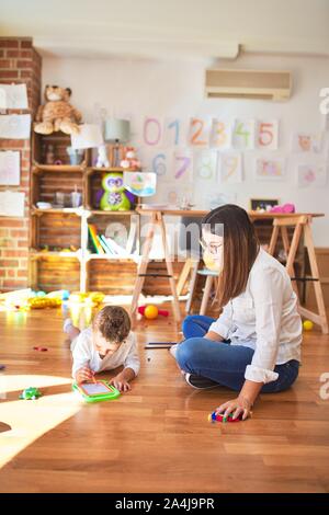 Beautiful teacher and toddler drawing using digital board around lots of toys at kindergarten Stock Photo