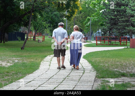 Middle aged couple is walking in forest. Trees on sides of trail. Elderly couple is walking in green park on holiday. Sunny summer day. Stock Photo