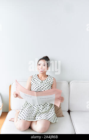 Young woman reading a newspaper sitting on a couch in the living room Stock Photo