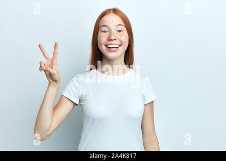 Attractive positive girl with freckles is showing peace sign and smiling. . Isolated on white background and copy space. close up portrait, body langu