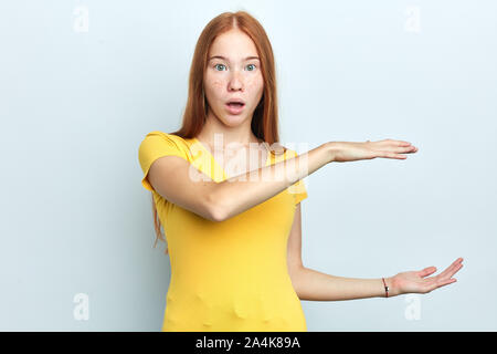 Shocked young surprised woman with wide open mouth shows something big in size with hands, demonstrates height of statue, isolated over white backgrou Stock Photo