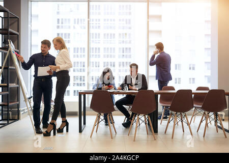 man explaining his ideas while standing next to flipchart while their colleagues sitting at the table discussing a problem, one man standing in front Stock Photo