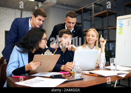 thoughtful pensive guy looking up trying to remember some information while sitting at the table in front of the screen of laptop with his colleagues. Stock Photo