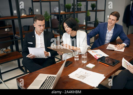 brunette woman holding folder in hands making notes explaining something to colleague while he is sitting at desk with serious expression and looking Stock Photo
