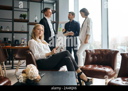 confident beautiul female leader of team is sitting on the armchair with crossed arms looking at the camera. three business people discussing the proj Stock Photo