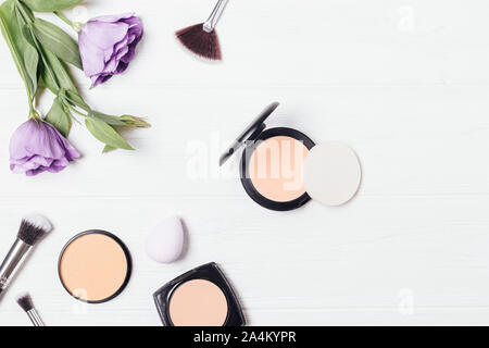 Compact powder and sponge applicator next to brushes, cosmetic products and fresh purple lisianthus on white background, flat lay composition. Stock Photo