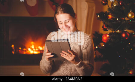 Toned portrait of happy smiling woman holding and using digital tablet on Christmas eve next to the fireplace Stock Photo
