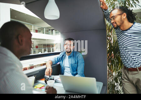 Smiling group of diverse creative professionals discussing a project together while working in an office meeting pod Stock Photo