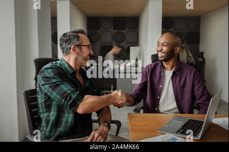 Entrepreneurs shaking hands in modern coworking workspace, people working in background Stock Photo