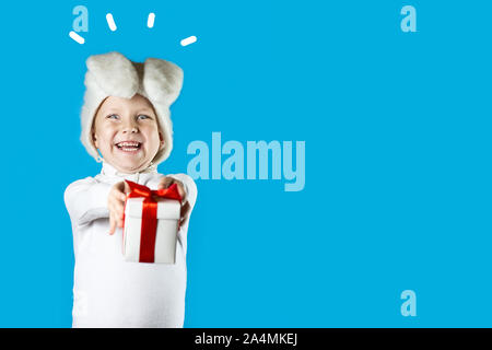 a cheerful boy in a white hare costume gives a gift with a red ribbon on a blue background Stock Photo