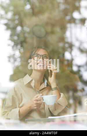 Young smiling businesswoman sitting behind the window of the cafe drinking coffee and talking on mobile phone Stock Photo