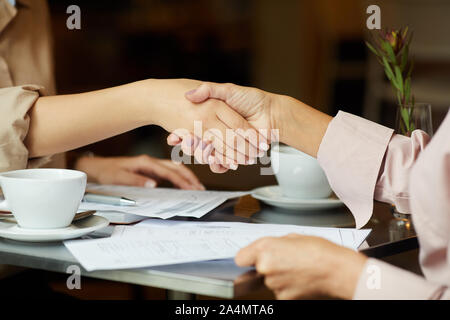 Close-up of two businesswomen shaking hands to each other while sitting at the table with cups of coffee during meeting in cafe Stock Photo