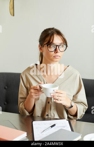 Young businesswoman in eyeglasses sitting at the table with documents thinking about something while drinking coffee Stock Photo