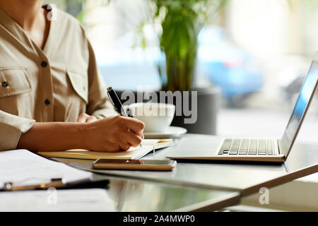 Close-up of woman sitting at the table in front of laptop and making notes in her notepad while drinking coffee Stock Photo