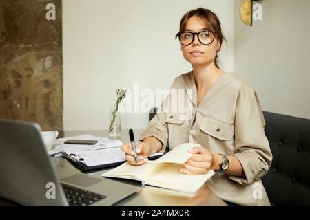 Serious young businesswoman in eyeglasses making notes in her notepad while sitting at the table with laptop at office Stock Photo