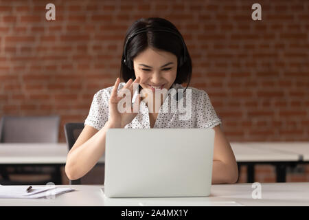 Smiling Asian businesswoman in headset waving hand, using laptop Stock Photo