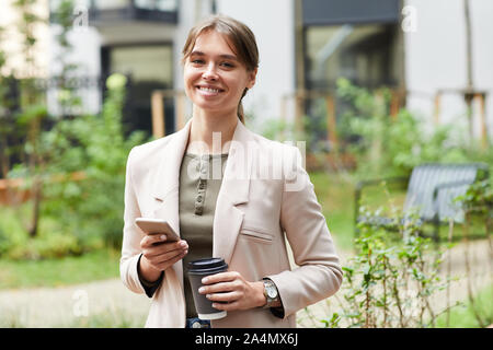 Portrait of young businesswoman smiling at camera while drinking coffee and using her mobile phone in the city Stock Photo