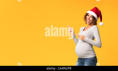 Joyful black pregnant woman in Santa hat pointing aside