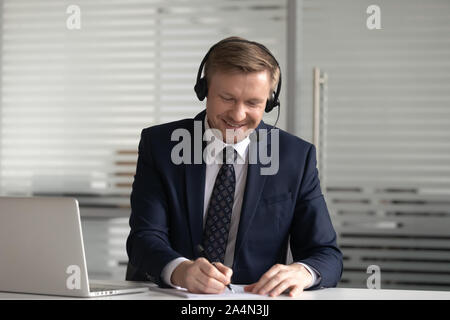 Smiling businessman in headset using laptop in office, making notes Stock Photo