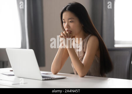 Thoughtful Asian businesswoman looking at laptop screen, pondering task Stock Photo