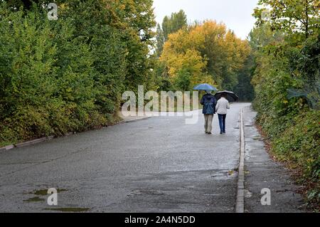 Rear view of an elderly couple with umbrella walking in the rain down an empty country road, Shepperton Surrey England UK Stock Photo