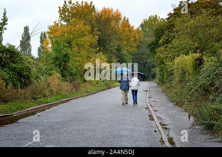 Rear view of an elderly couple with umbrella walking in the rain down an empty country road, Shepperton Surrey England UK Stock Photo