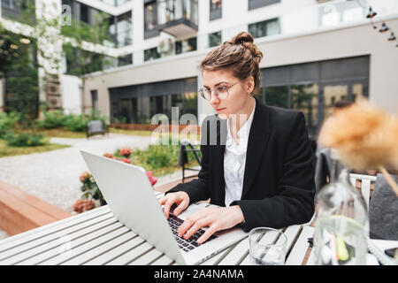 Young business woman sitting at coffee shop on veranda and working on laptop. Portrait of female freelancer wearing glasses with computer Stock Photo