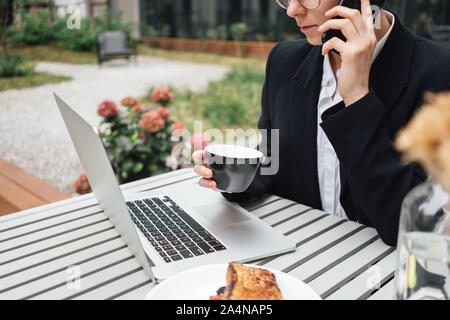 Beautiful business woman sitting on veranda at cafe while working on laptop and talking on smartphone with client. Young female freelancer have conver Stock Photo