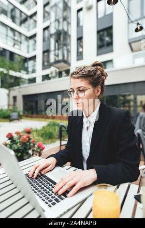 Portrait of female freelancer wearing glasses with computer. Young business woman sitting at coffee shop on veranda and working on laptop Stock Photo