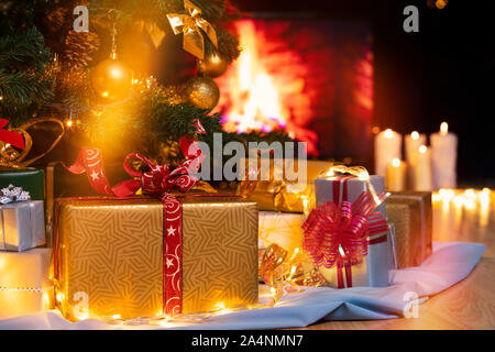Stack of packed gift boxes under Christmas tree against burning fireplace. Lots of Christmas gifts under the tree. Candles on wooden floor. Focus on g Stock Photo