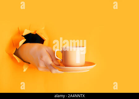 Hand holding a cup of coffee through a hole in torn saffron or light orange paper wall. Coffee break concept, minimalism Stock Photo