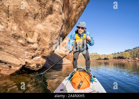 senior male is paddling stand up paddleboard along sandstone cliff on Horsetooth Reservoir in northern Colorado Stock Photo