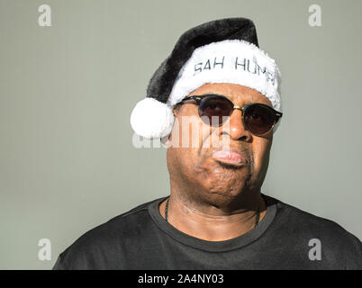 a frowning middle aged African American man wearing a black and white Santa Claus hat saying Bah Humbug on it against a solid background Stock Photo
