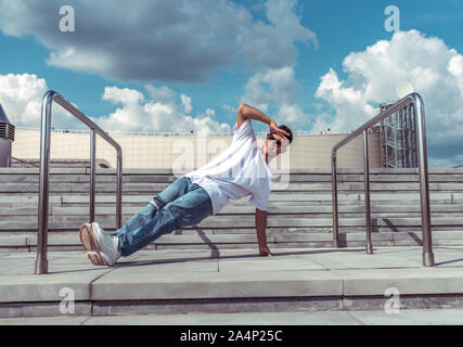 dancer on one arm dances break dance, hip-hop. In summer, city background steps clouds. Active youth lifestyle, young male dancer, fitness movement Stock Photo