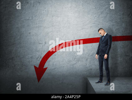 Upset businessman standing on the edge of abyss with red arrow going down Stock Photo