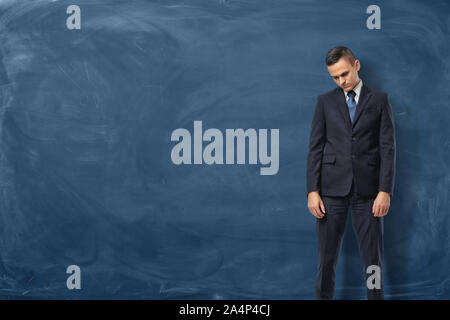 Sad businessman in suit standing with bowed head and arms down near the blue chalkboard Stock Photo