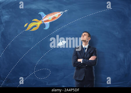 A businessman standing with his arms folded on the dark blue background, he is looking up at the pictures of the rocket and the UFO drawn on the wall. Stock Photo