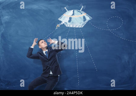 Businessman frightened by flying saucer painted on blue blackboard behind him Stock Photo