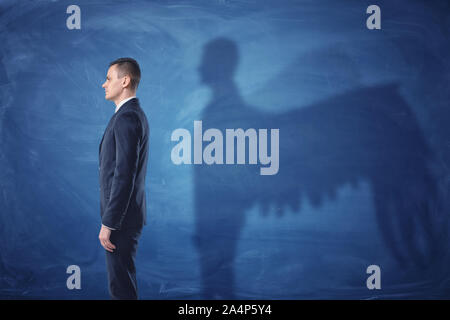 Businessman is standing in profile and casting a shadow of angel wings on blue chalkboard background Stock Photo