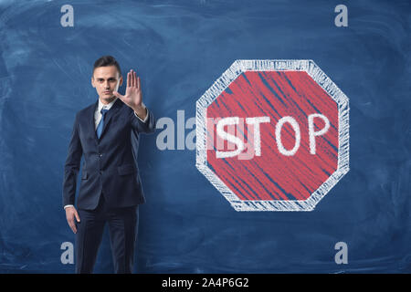 Businessman standing with his arm forward and traffic-sign 'Stop' painted on blackboard behind him Stock Photo